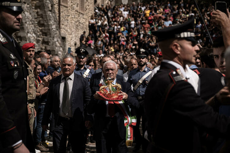 Two men lead a procession of people between rows of Italian Carabinieri and Soldiers. One is holding a large crown and wearing a sash of the colors of the flag of Italy.