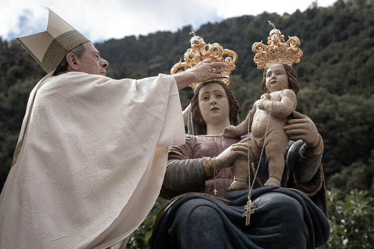 A close-up of a priest placing a golden crown on the statue of the Madonna and Child against a forest background.