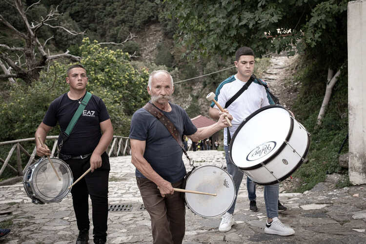 An older man and two younger men play drums outside on a mountain trail.