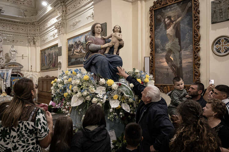 Inside a church the statue of the Madonna and Child is surrounded by flowers, and a group of people in the foreground reach up to touch the statue's knees.